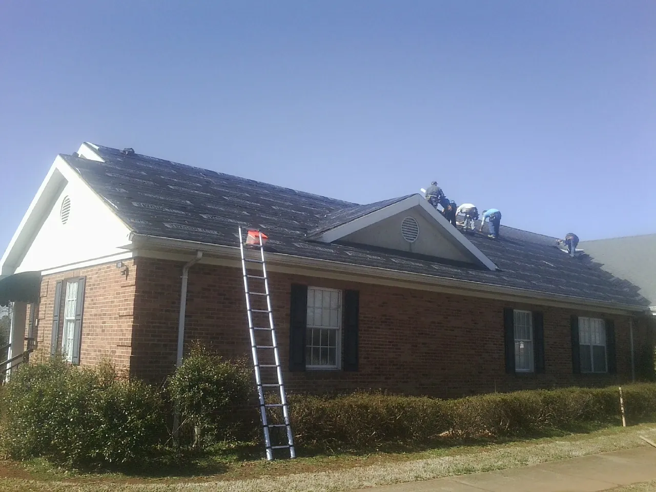 Crew tearing shingles off a brick ranch roof with a ladder set — Emergency Roof Repair work by our crew in Kokomo