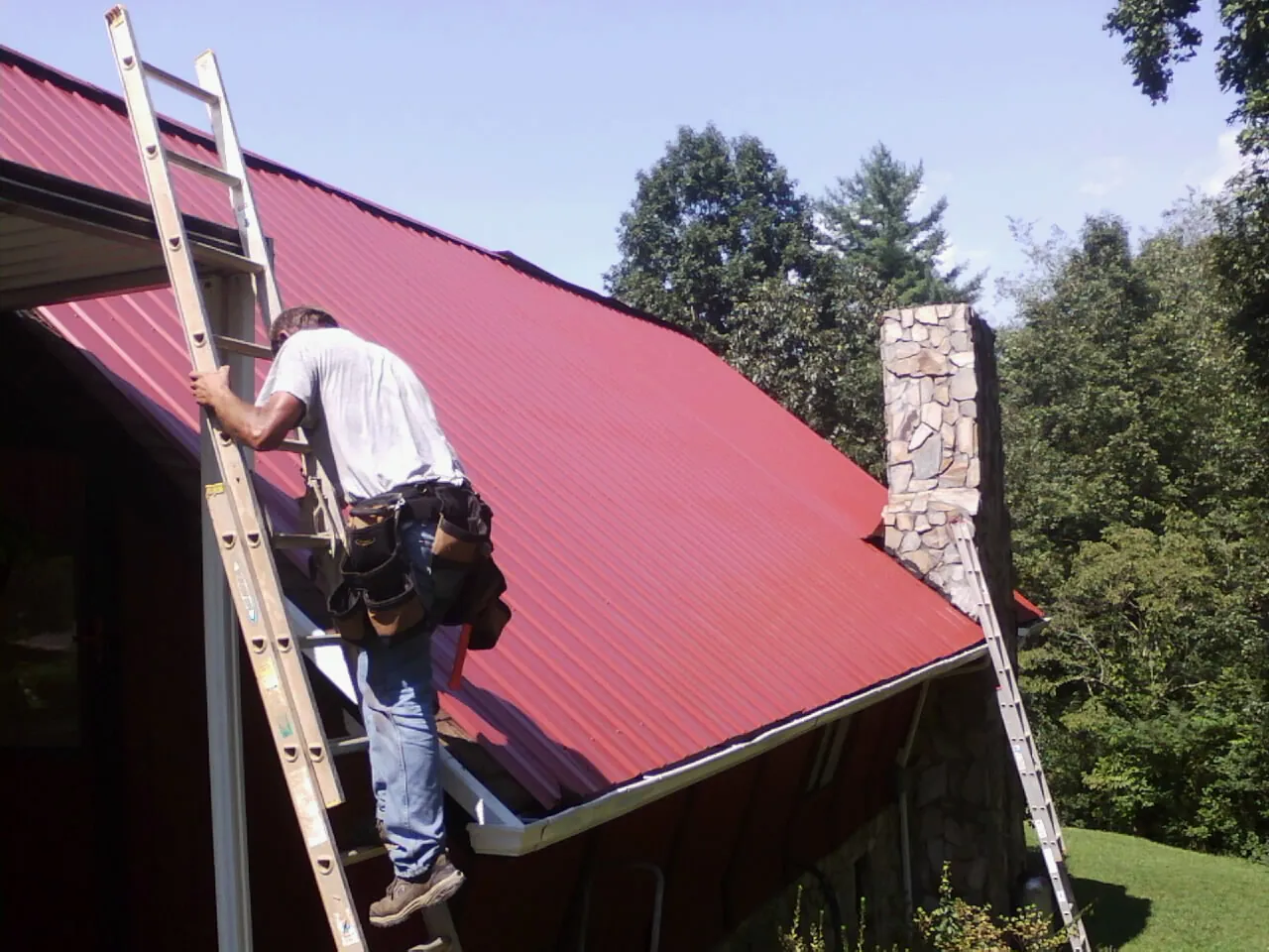 Roofer on a ladder beside a red metal roof and stone chimney — Soffit & Fascia Repair by Roofing Brothers in Kokomo